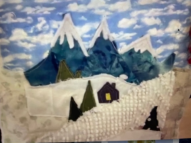 fabric art formed into a winter scene with snow covered mountains and log cabin